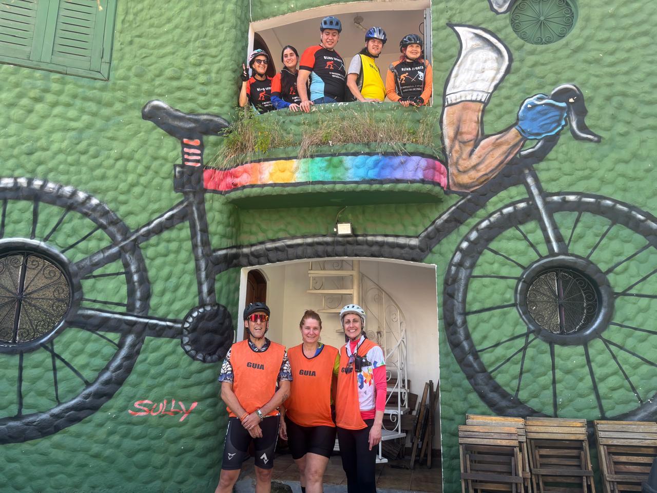 Fachada de prédio verde com textura, adornada por um grande grafite de bicicleta preta, ocupando quase toda a parede. As rodas incorporam janelas arredondadas e, na parte superior, um grafite de pernas pedalando. No topo, seis pessoas com roupas de ciclismo e capacetes estão em um balcão, sorrindo e olhando para a câmera. Abaixo, em frente à porta, três pessoas (duas vestindo coletes laranja escritos “GUIA” e uma de camisa clara com detalhes laranja e rosa, todos também com capacetes de ciclismo) posam para a foto. À direita, pilha de cadeiras de madeira encostadas na parede. Ambiente descontraído e alegre, transmitindo espírito de grupo e aventura.